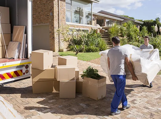 Mover carrying a cardboard box outdoors with packed items nearby.