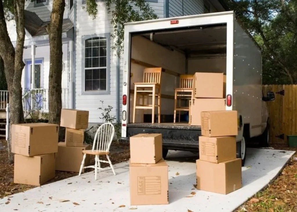 A moving truck parked outside a white house with cardboard boxes and chairs being loaded in Edmond, OK.